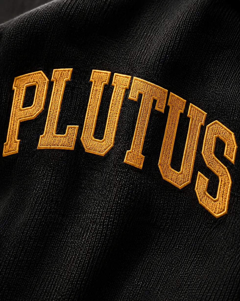 Close detail of the arched PLUTUS lettering embroidered in gold on black knit.