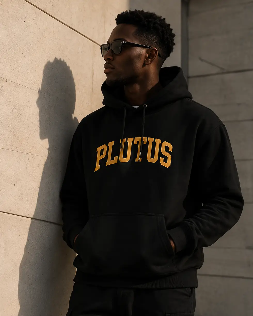 A man wearing the black PLUTUS hoodie against a concrete wall in daylight.